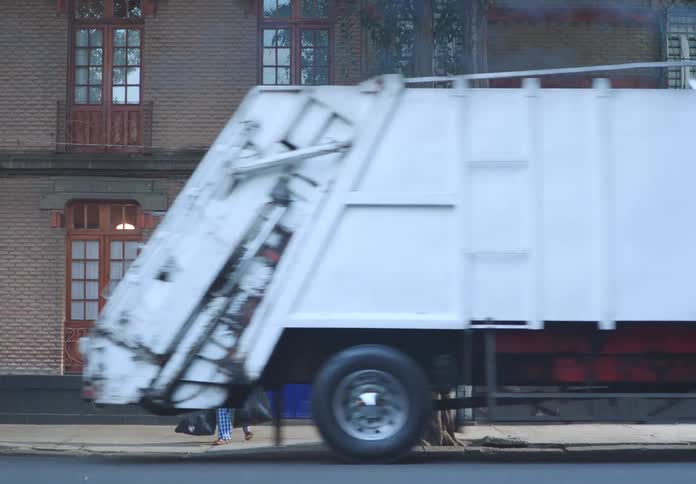 A man carrying garbage bags in both hands runs after a moving garbage truck that does not stop