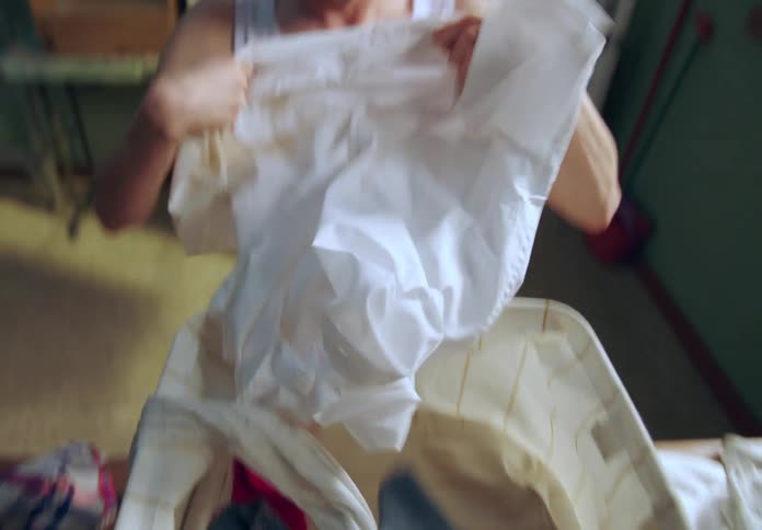 Man hastily searching inside a basket of clothes finds a shirt smells it but probably stinks and makes a bad face and leaves