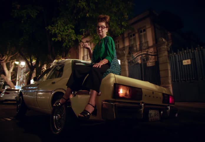 A woman wearing eyeglasses is sitting on the trunk of a dodge car while playing with a fruit