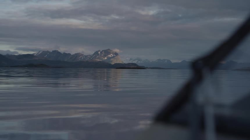 A calm body of water with mountains in the distance under a cloudy sky as seen from a boat