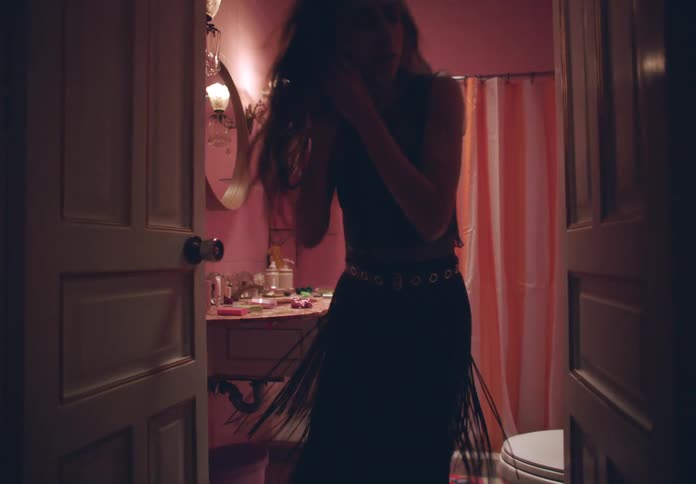 Woman in a black dress leans over a sink in a bathroom putting on makeup and then runs down the hallway of the house
