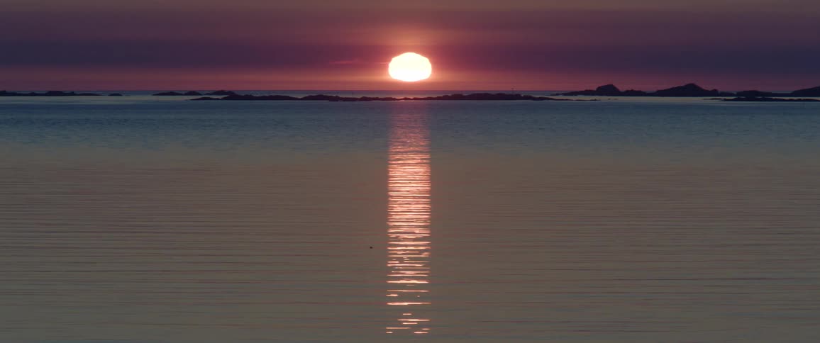 A sunset over a calm sea reflects a shimmering path of light towards the viewer with dark landmasses visible on the horizon