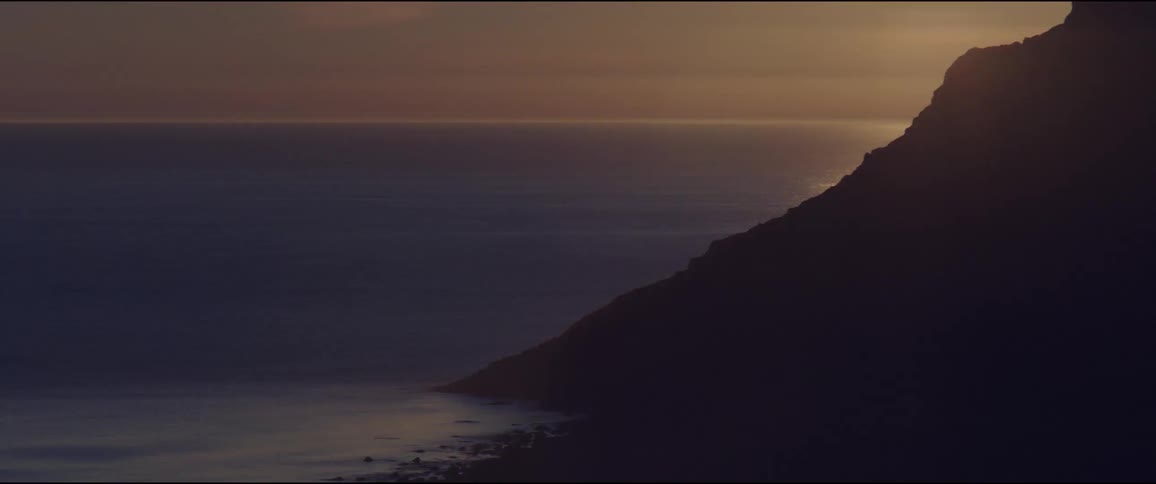 From an elevated vantage the coastline appears in partial silhouette against the oceans warm evening glow where the water meets a gently fading horizon under soft light