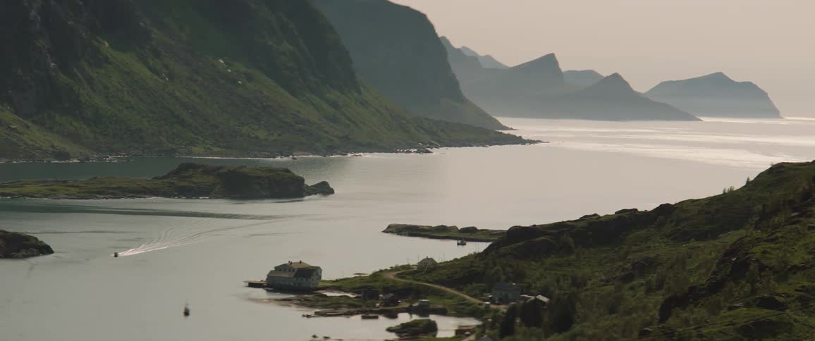 From a lofty vantage a small coastal town borders a narrow canal where boats pass through calm waters while towering green cliffs and rugged silhouettes frame the background under a soft sunlit haze