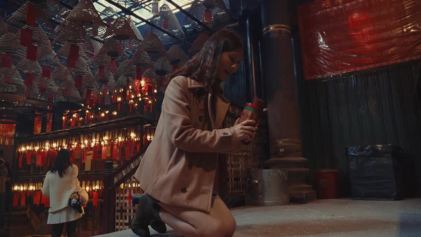 A woman kneels in an asian temple shaking a container until an incense stick falls out then smiles and begins to pray