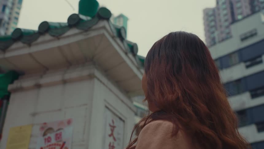 A woman with long brown hair walks away from the camera towards a building with a traditional asianstyle roof