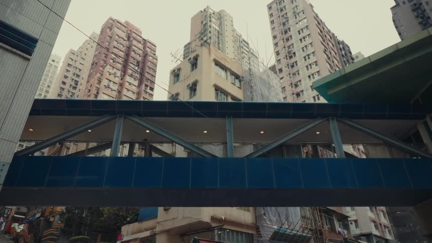 An elevated pedestrian bridge spans a busy hong kong with individuals seen going across its expanse set against a backdrop that is including tightlypacked highrise urban structures and a hazy sky