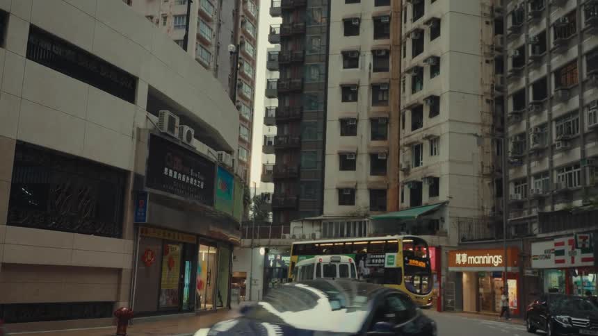 A street scene in hong kong shows a mix of vehicles including a taxi and a doubledecker bus moving through a narrow street lined with tall densely packed buildings