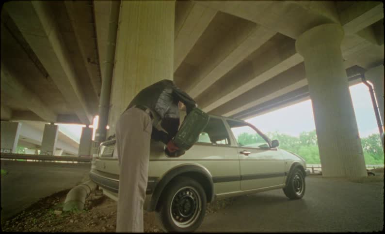Man empties the gas from a gas can into a car parked under a raised highway and then throws it away the can