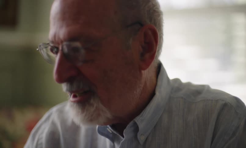 A closeup shows a mature man with a beard and glasses talking in a room