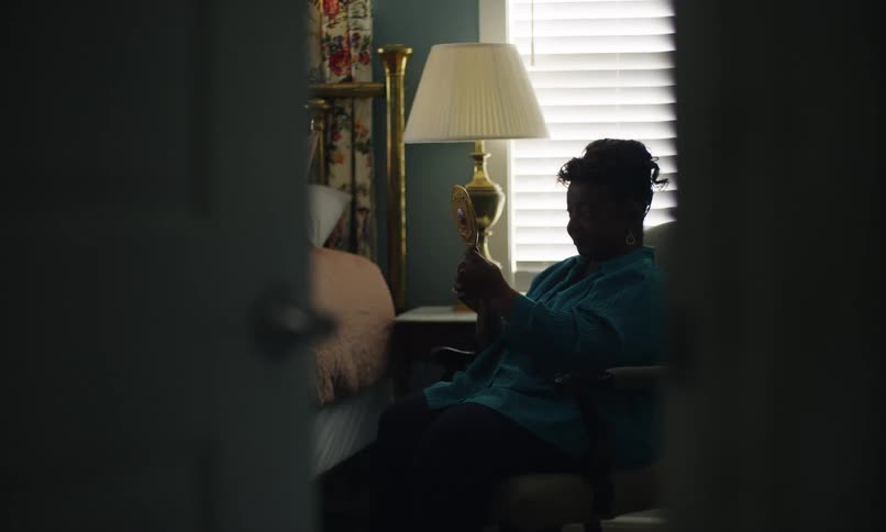 The elderly woman is sitting in the hair and she is holing a handheld mirror and putting on makeup