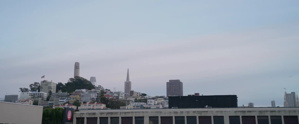 San franciscos cityscape marked by the iconic coit tower and transamerica pyramid is on display amidst a hazy and pale urban expanse as it is under a white light blue sky with the camera panning slightly to the right