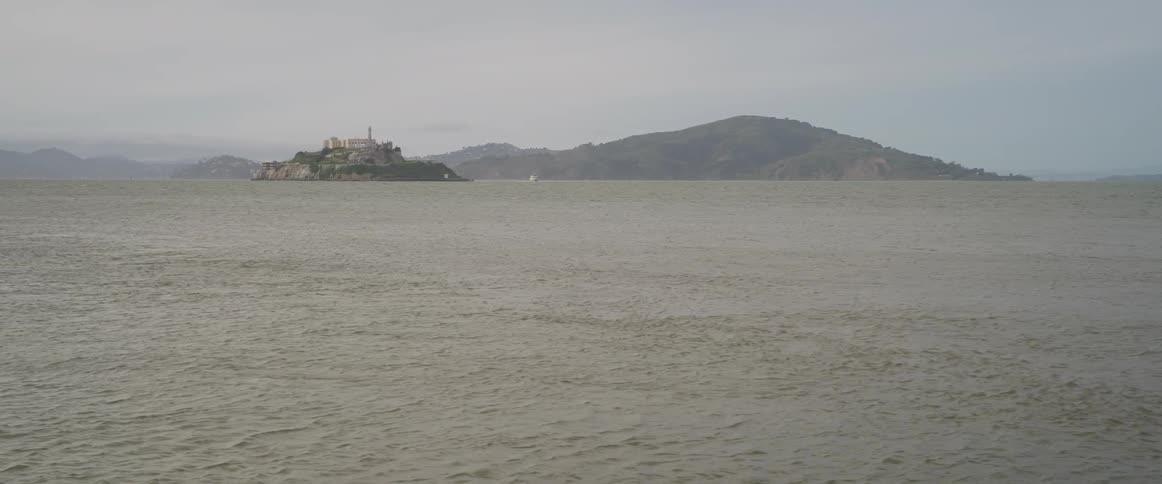 A view across the expansive choppy waters of san francisco bay towards the distant silhouette with the infamous alcatraz island with the old buildings set beneath an overcast pale gray sky