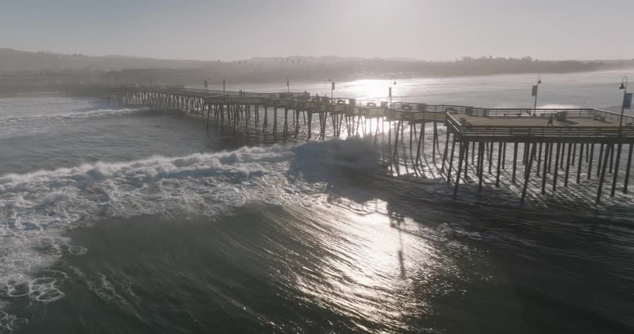 An aerial view shows a long pier extending into the ocean with large waves crashing against it and the shoreline under a hazy sky