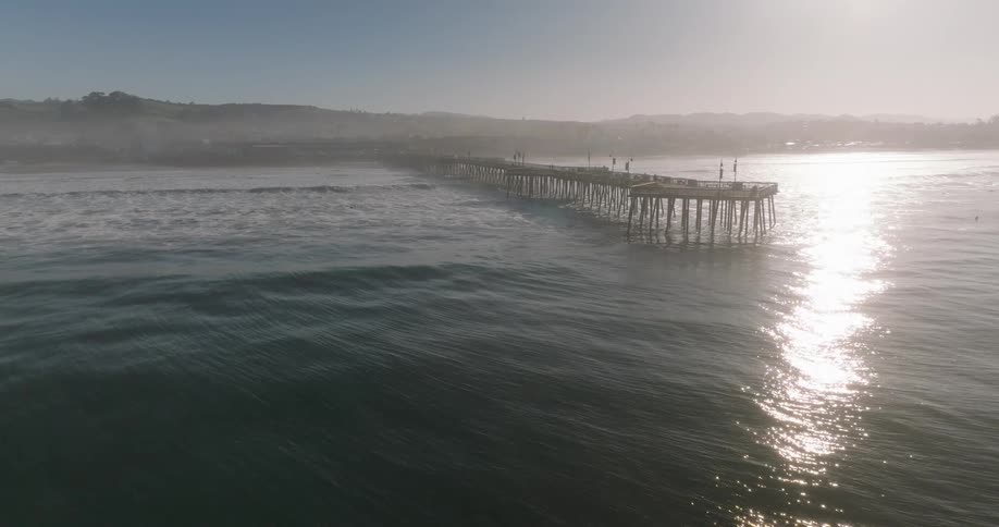 An aerial view of a long pier extending into the ocean with waves crashing against its sides and a town visible in the background under a bright sky