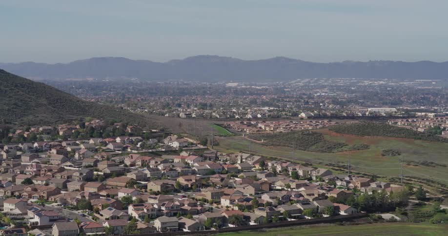 Aerial view of a densely packed suburban neighborhood with houses clustered together extending towards a distant cityscape and mountains under a hazy sky