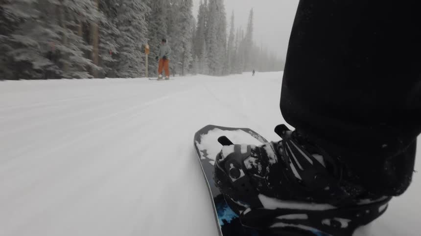 Closeup of a persons legs and a man in the background skiing down a snowy mountain at high speed