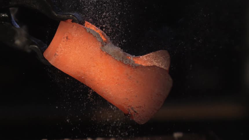 White dust cascades onto hot metal held with tongs in a dimly lit blacksmith workshop