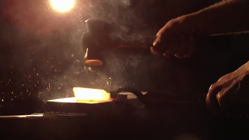 A blacksmiths hands work a glowing hot piece of metal with a hammer on an anvil sending sparks and smoke into the air