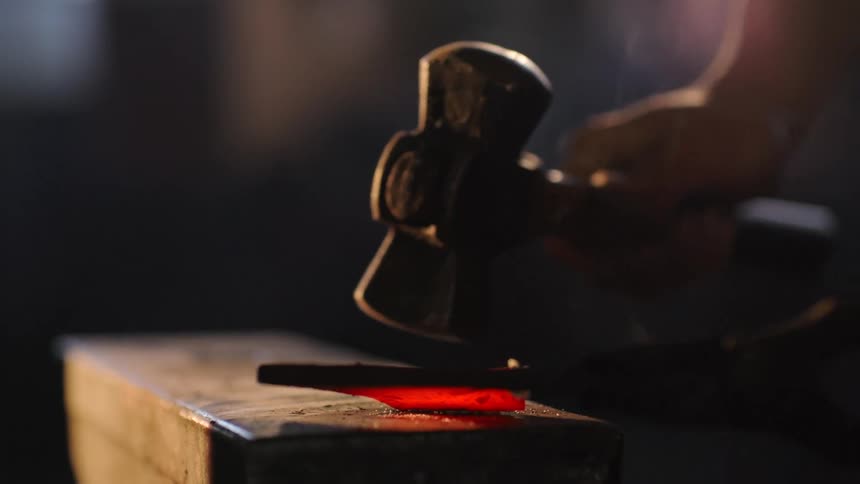 A hammer strikes a glowing piece of redhot metal on an anvil while being steadied with tongs