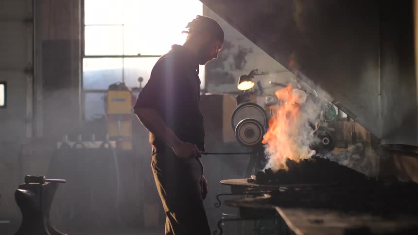 A blacksmith works at a coal fire forge in a brightly sunlit workshop near a large window