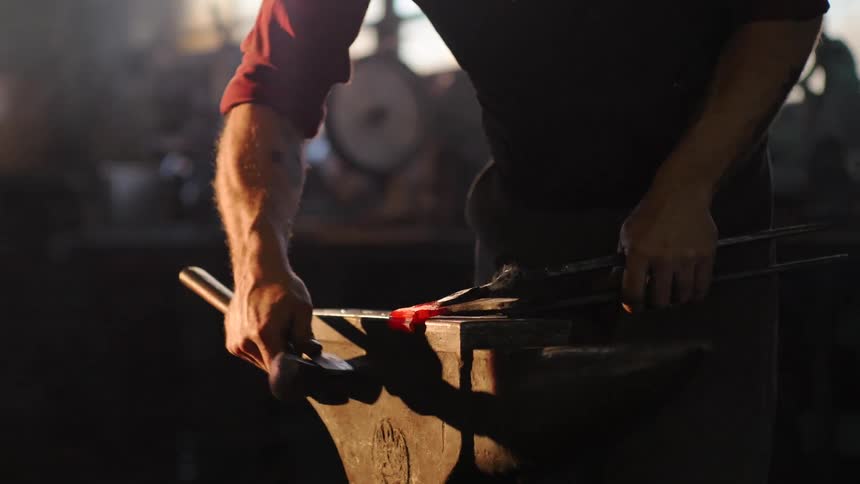 A blacksmith shapes glowing hot metal on an anvil with a hammer and tongs in a dimly lit workshop