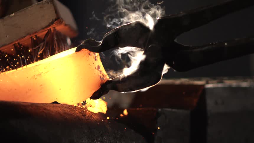 A pair of blacksmith tongs firmly grip a glowing molten hot piece of metal with sparks and steam rising as intense heat radiates from the forging process