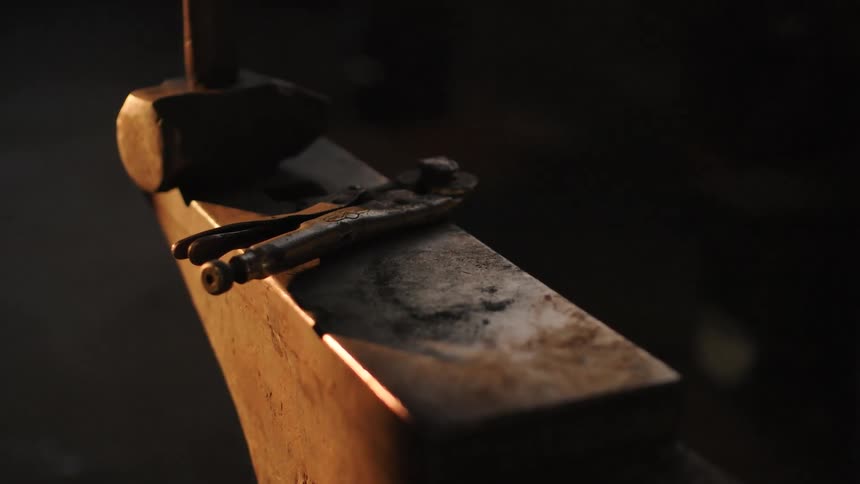 A closeup shows a hammer and a wrench resting on an anvil in a dimly lit workshop