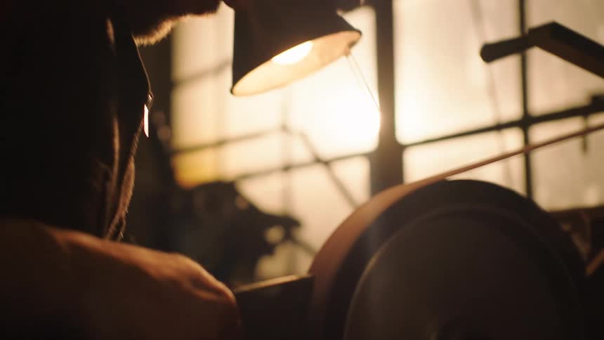 A closeup shows a man wearing safety glasses sharpening an axe on a spinning wheel in a workshop with bright sparks flying
