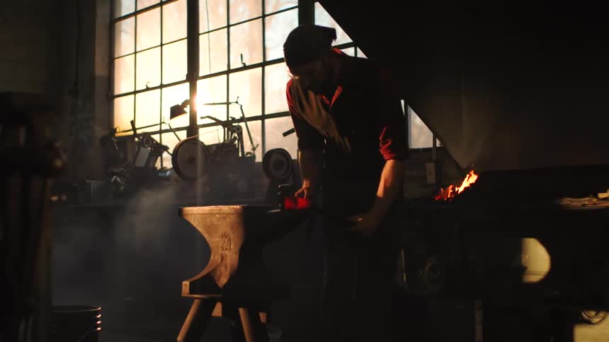 In a dimly lit workshop with sunlight streaming through the windows a blacksmith works at an anvil hammering a piece of red hot metal near a fiery forge