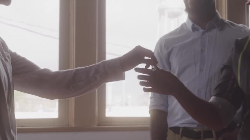 A man hands a key to a smiling woman in a brightly sunlit room with large windows