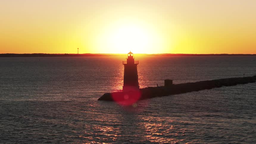 An aerial view slowly moves away from a silhouetted lighthouse on a rocky outcrop set against a vibrant sunset over the ocean
