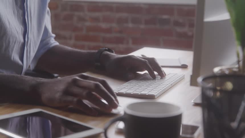 A man with a light blue shirt types on a white keyboard at a desk with a tablet and mug visible in the foreground