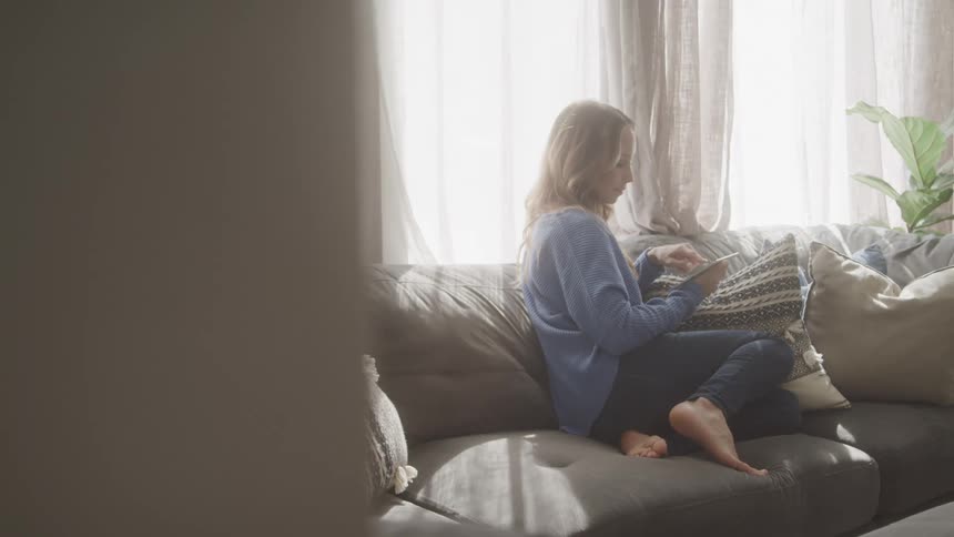 A woman sits on a couch in a sunlit living room near a bright window holding a tablet in her hand