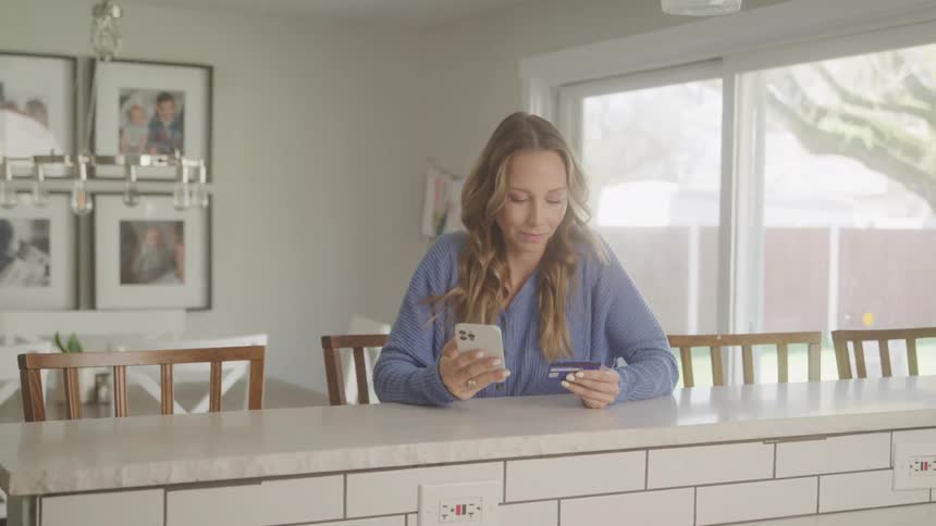 A blonde woman sits at her kitchen snack bar and enters credit card information into her smartphone