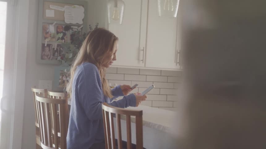 A blonde woman with long hair sits in a kitchen on a bright day holding a phone and a credit card
