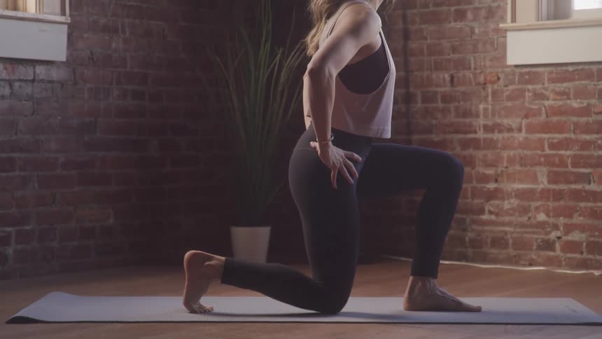 A woman with blonde hair performs lunges on a yoga mat in a room with exposed brick walls