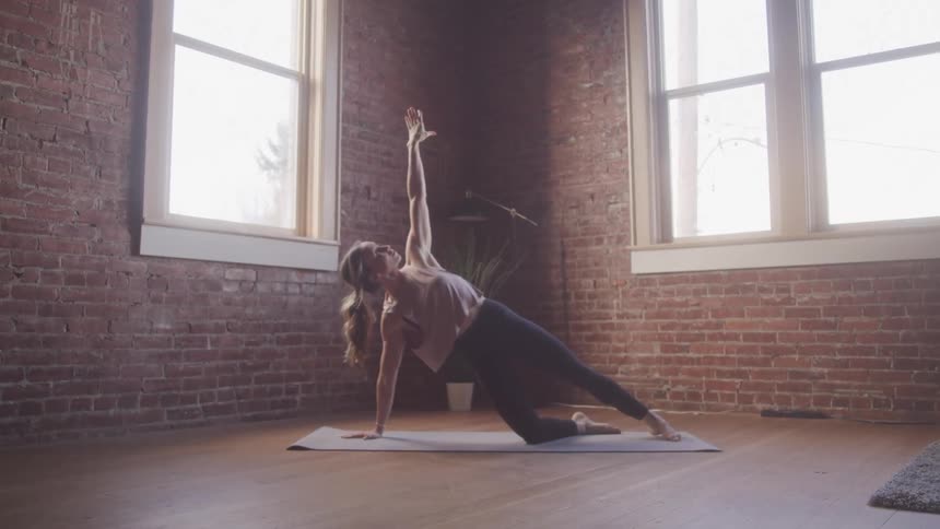 A woman stretches on a mat on the floor in a sunlit room with a brick wall and large windows