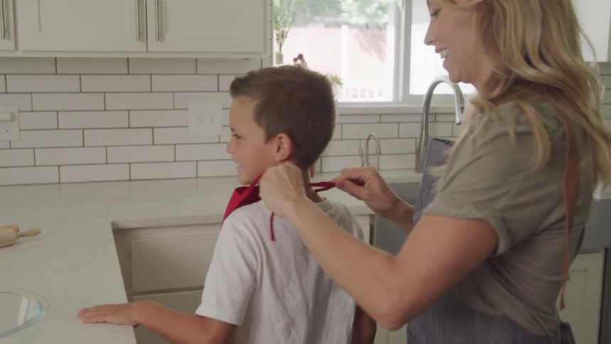 A blonde woman ties a red apron around her sons neck while standing in the kitchen