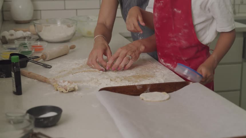 Child wearing a red apron puts the dough into a mold while another person helps her in the kitchen