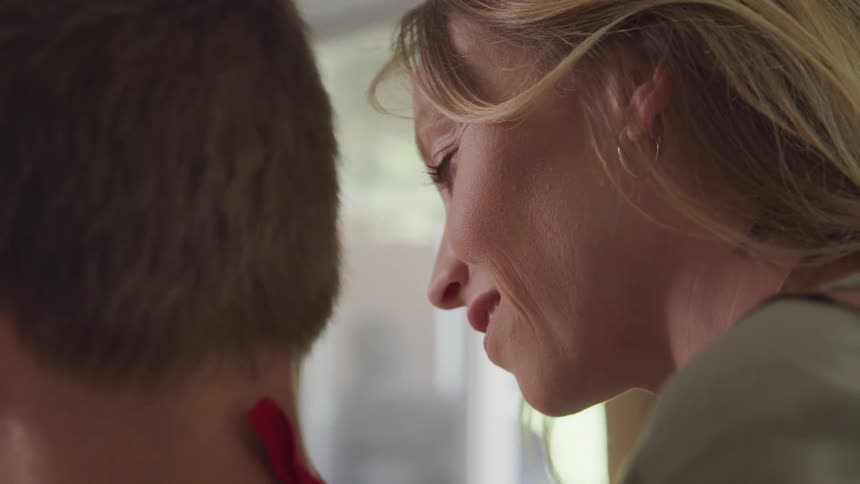 A mother with blonde hair in profile looking down next to her son with dark brown hair in the kitchen