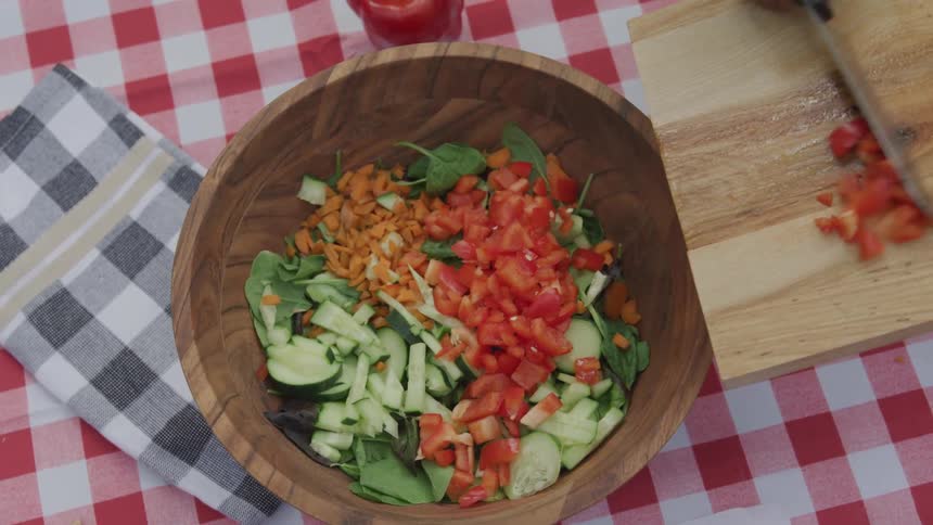 Chopped red bell pepper is scraped from a cutting board into a wooden bowl filled with other salad ingredients on a redcheckered tablecloth