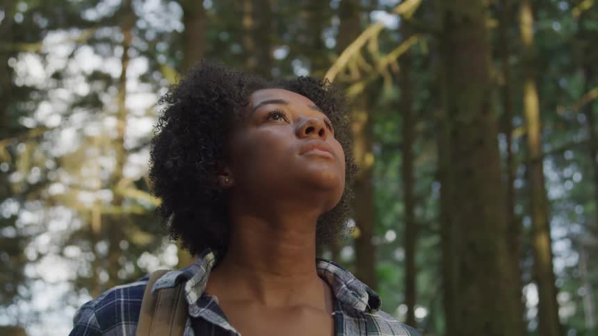 A woman with dark curly hair gazes upwards towards the sunlight filtering through tall trees in a forest