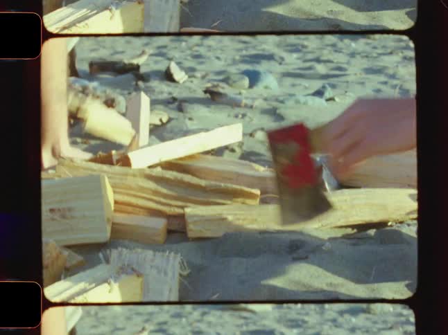 A young man with long blonde hair wearing sunglasses and a baseball cap chops wood near a campfire on the beach in the morning