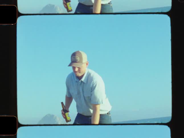 A man with a beer bottle is pulling driftwood towards a beach campfire in the morning