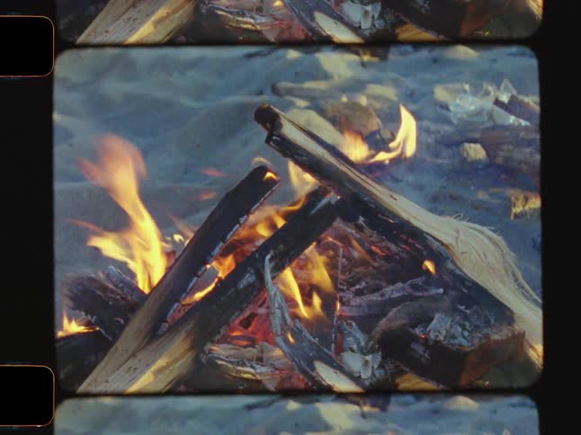 Flames rise from burning wooden logs in a campfire surrounded by ash and debris