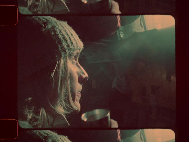 Blonde man drinks from a thermos while looking out a small window at dawn
