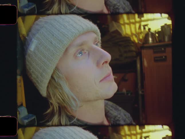 A closeup shows a young man with long hair and a beanie sitting by the open door of a van and looking upward