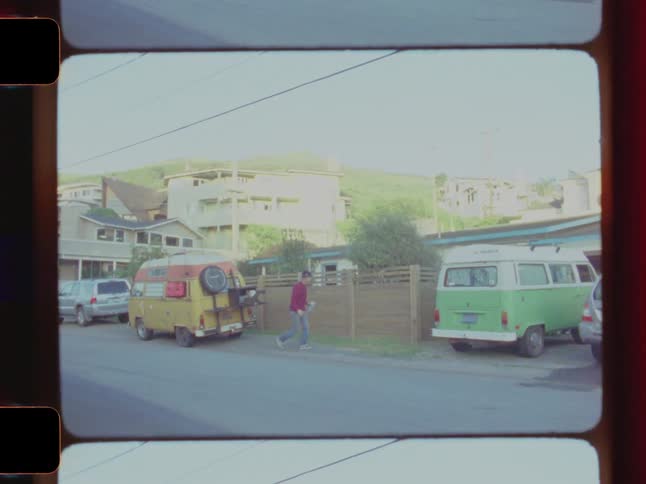 Man walks along a fence and passes a van parked near a building on a sunny day