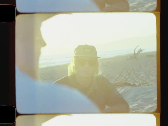 A man wearing sunglasses and a baseball cap sits on a sandy beach talking to another man under the sunlight